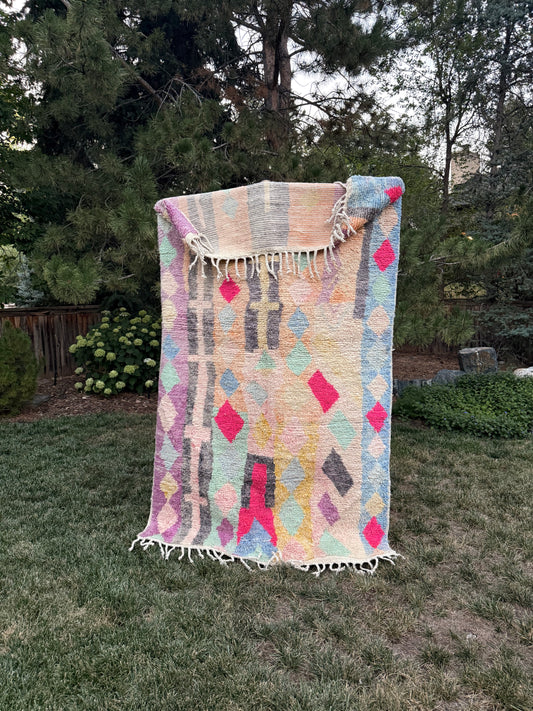 A woven Moroccan rug with fringe, featuring a pastel geometric pattern in pink, yellow, blue, and gray on a cream background, displayed outdoors on a mountain lawn.