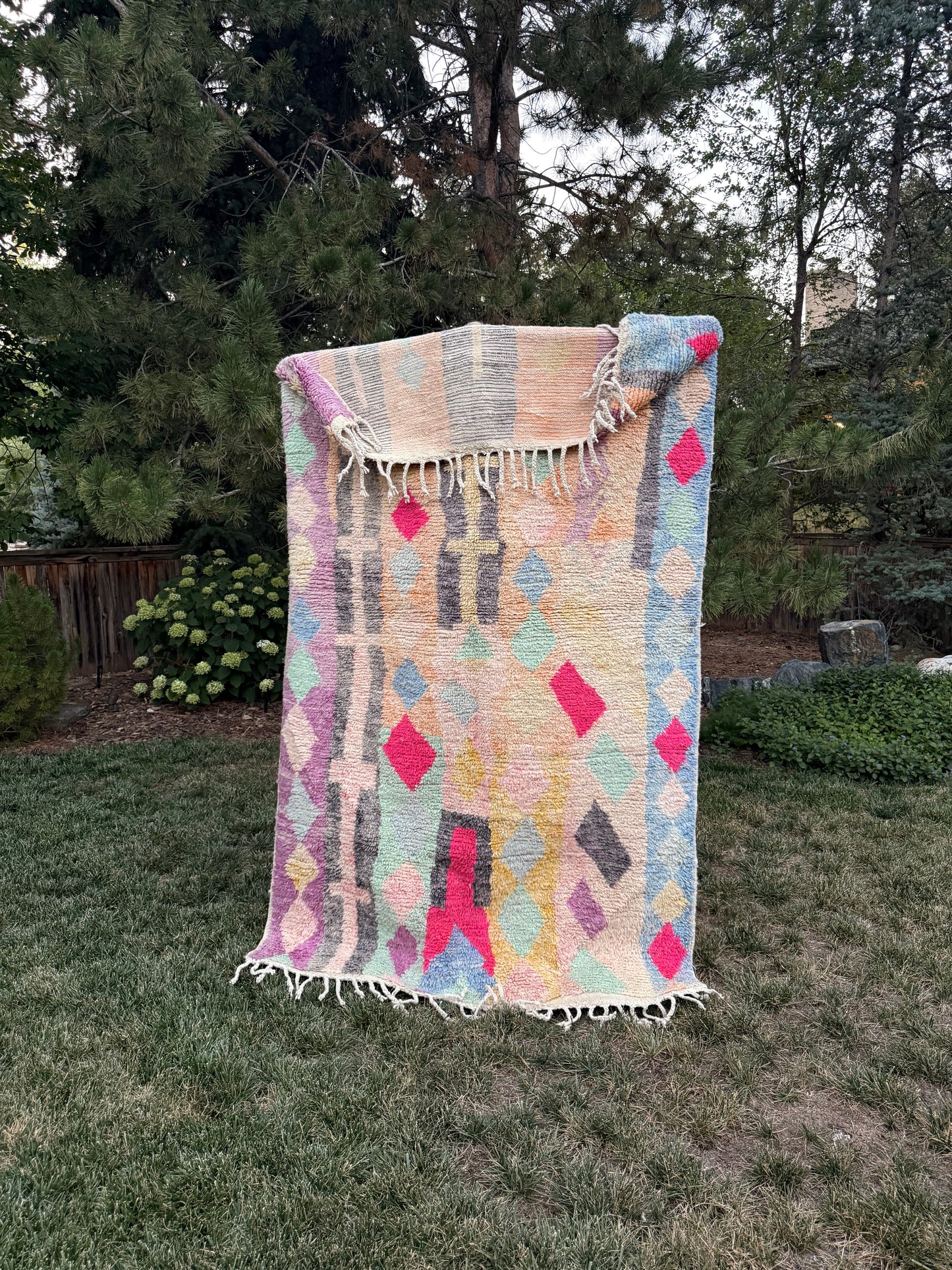 A woven Moroccan rug with fringe, featuring a pastel geometric pattern in pink, yellow, blue, and gray on a cream background, displayed outdoors on a mountain lawn.
