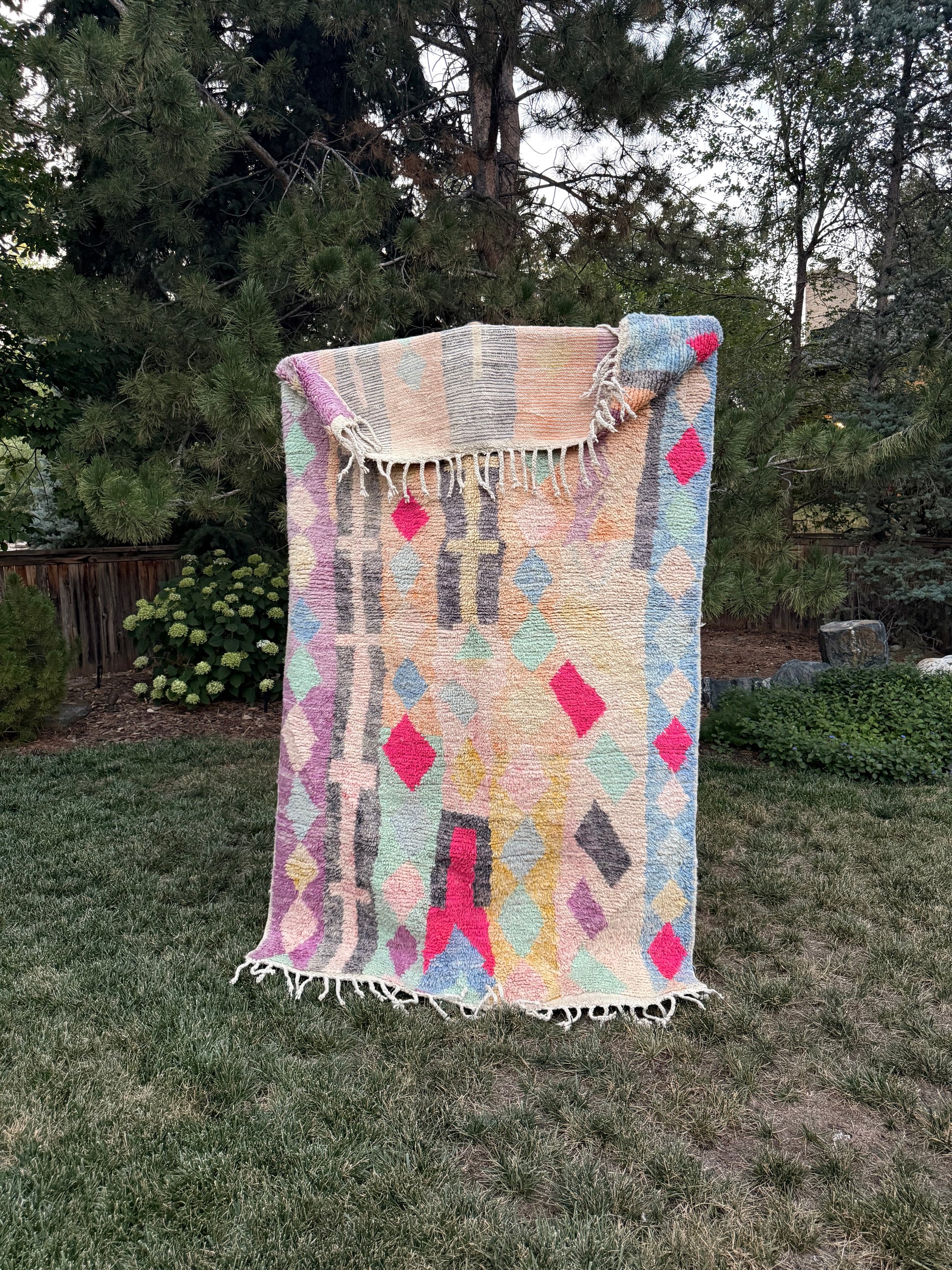 A woven Moroccan rug with fringe, featuring a pastel geometric pattern in pink, yellow, blue, and gray on a cream background, displayed outdoors on a mountain lawn.
