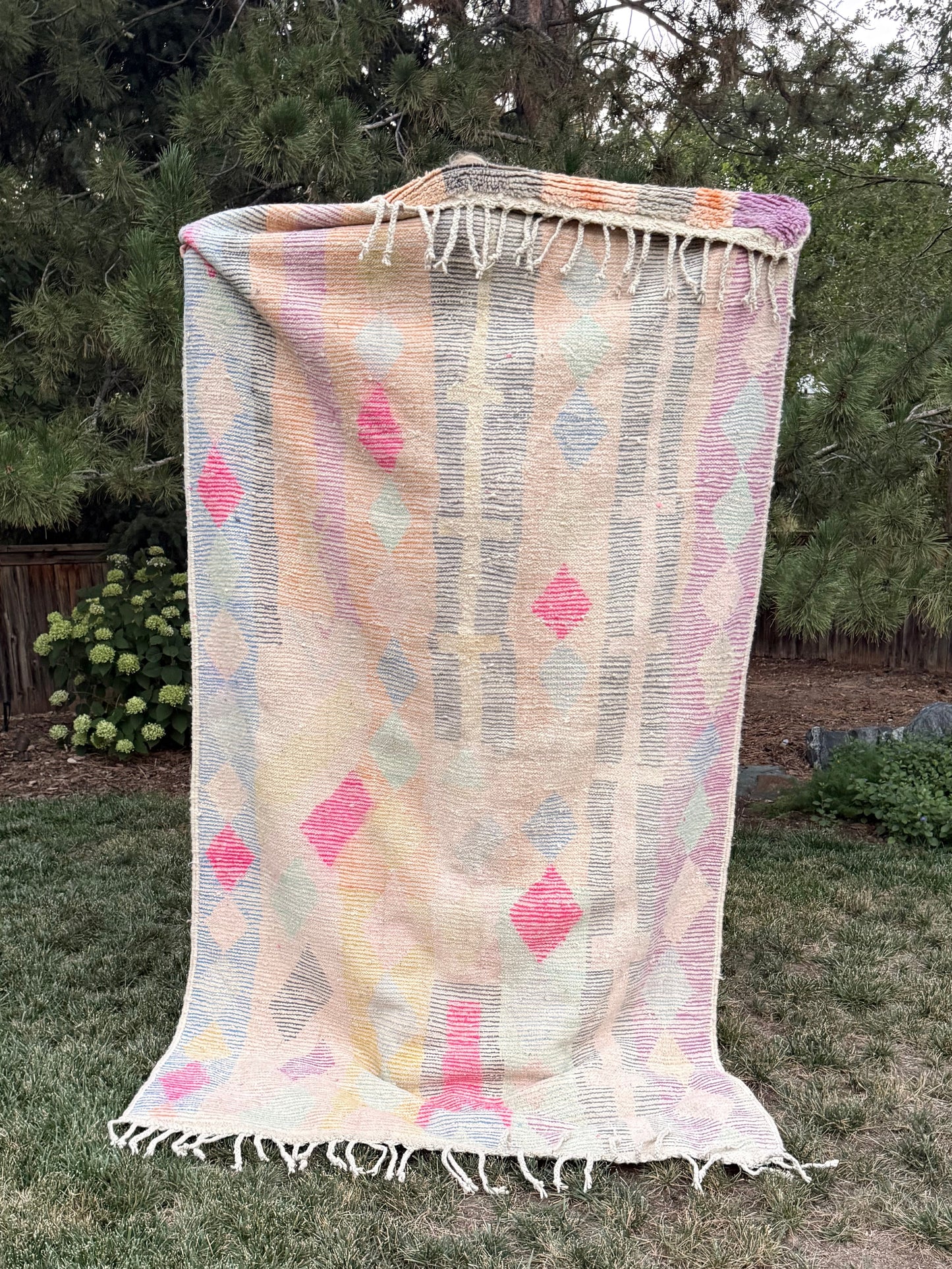 A woven Moroccan rug with fringe, featuring a pastel geometric pattern in pink, yellow, blue, and gray on a cream background, displayed outdoors in the mountains.
