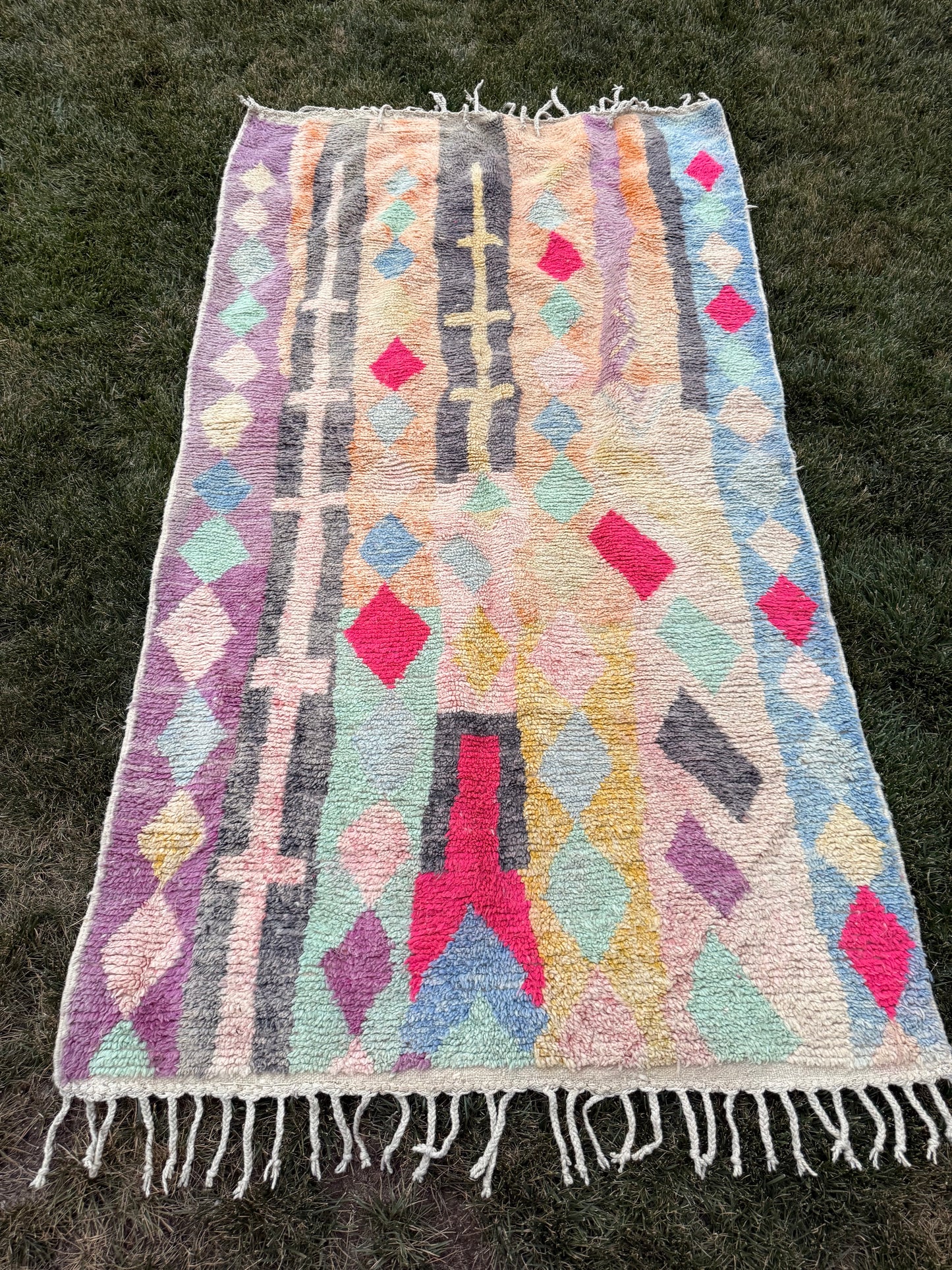 A rectangular woven rug with fringe, laid flat on green grass. The rug features a multicolor geometric pattern with vertical stripes and diamond motifs in pink, purple, blue, green, yellow, and gray on a cream base.