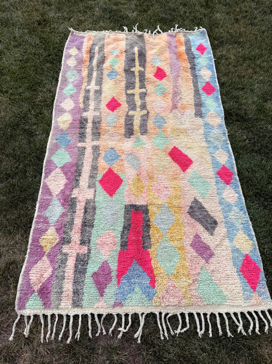 A rectangular woven rug with fringe, laid flat on green grass. The rug features a multicolor geometric pattern with vertical stripes and diamond motifs in pink, purple, blue, green, yellow, and gray on a cream base.